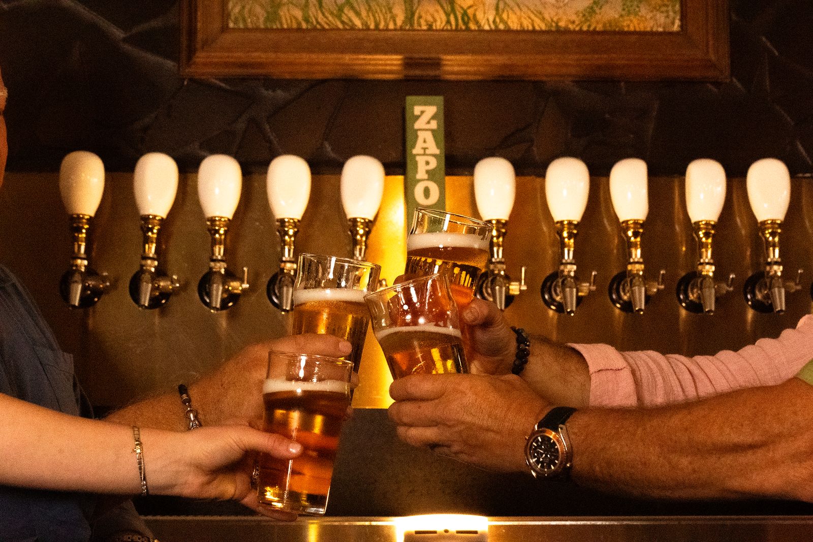 Four friends clinking glasses of ZAPO in front of a long row of brass taps marked by a green ZAPO tap handle.