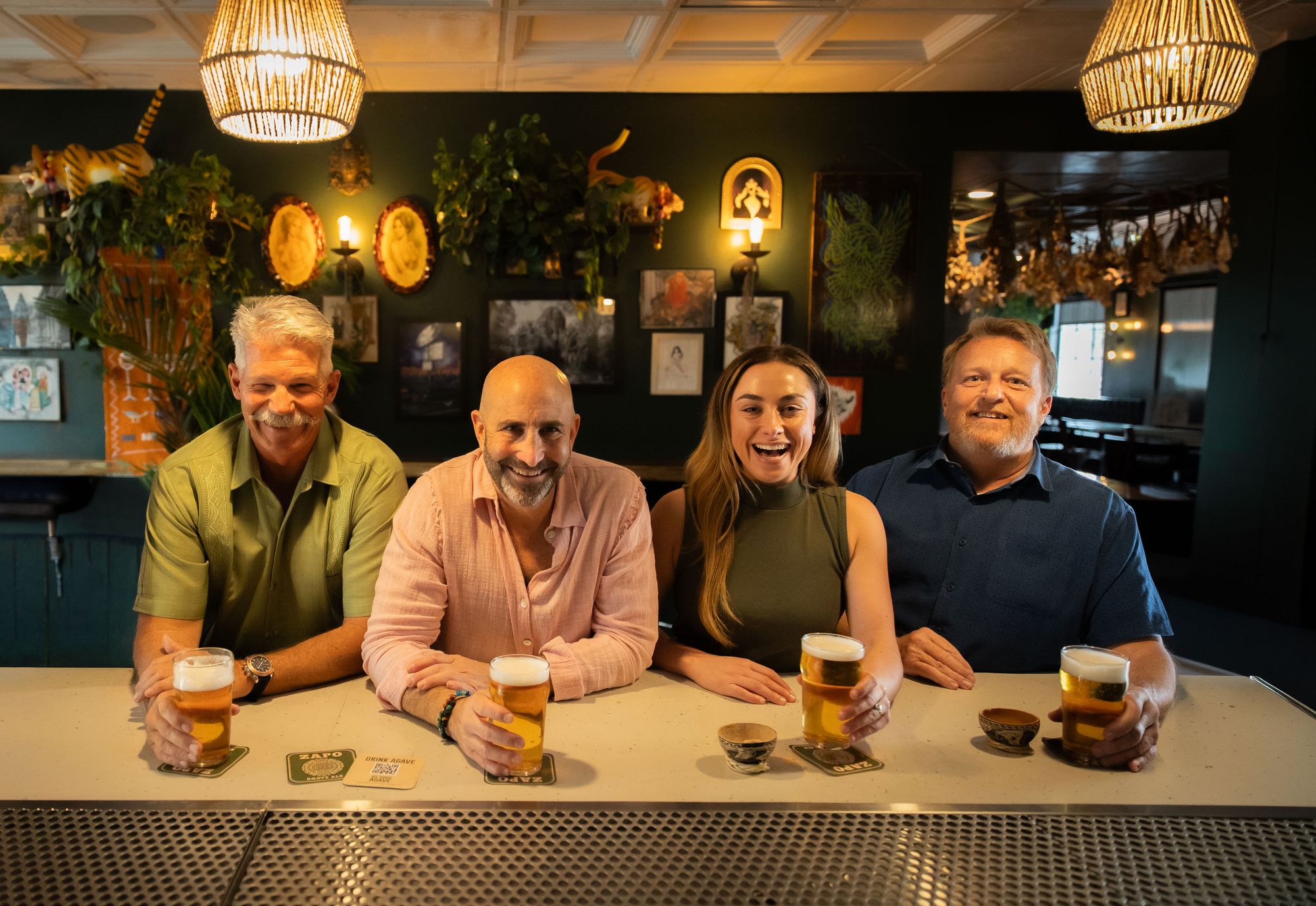 The four All Things Agave team members smiling behind a bar, each holding a pint of ZAPO on branded coasters.